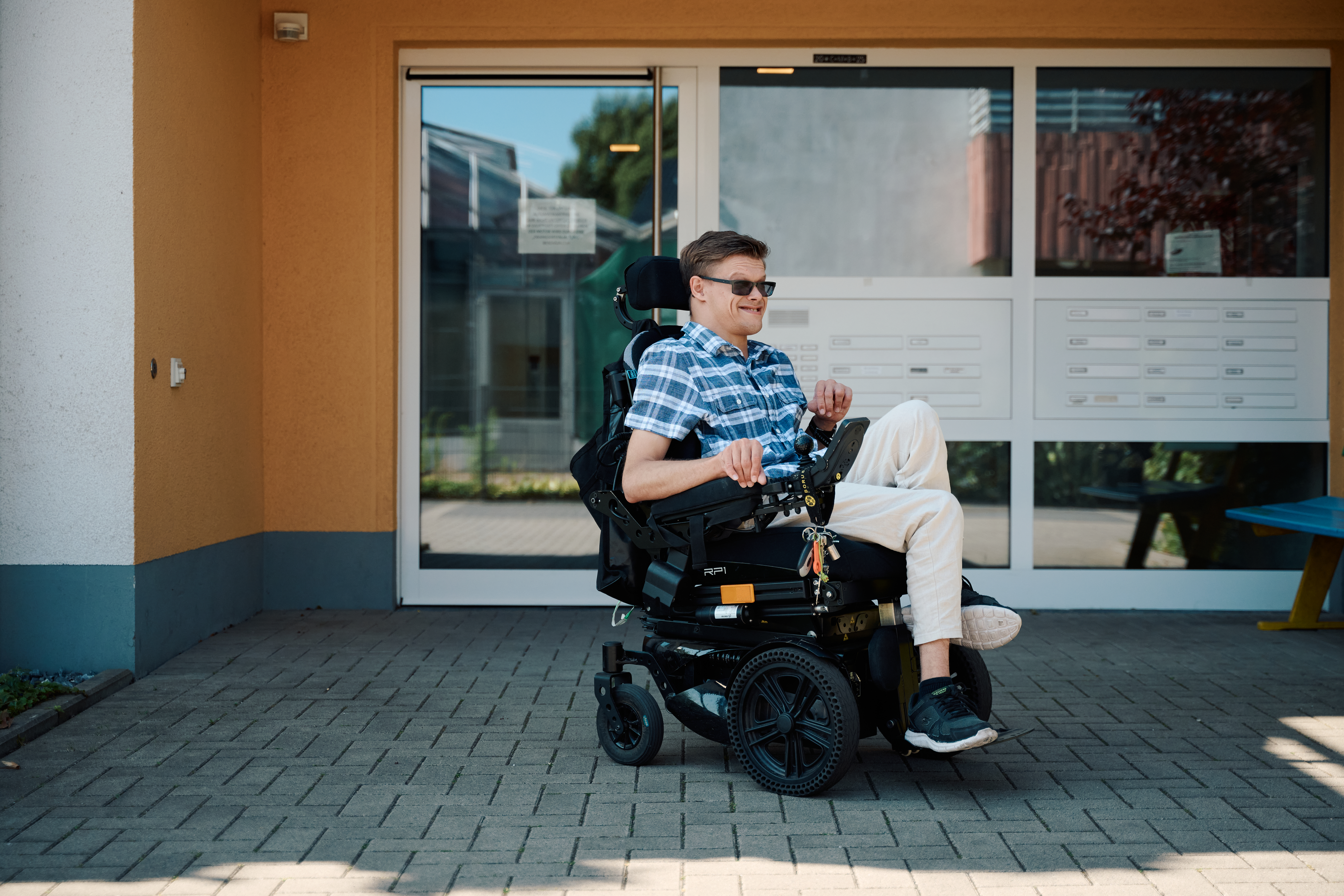 Ein junger Mann im sitzt im elektrischen Rollstuhl vor dem Eingangsbereich eines Mehrfamilienhauses. Ein junger Mann im sitzt im elektrischen Rollstuhl vor dem Eingangsbereich eines Mehrfamilienhauses.