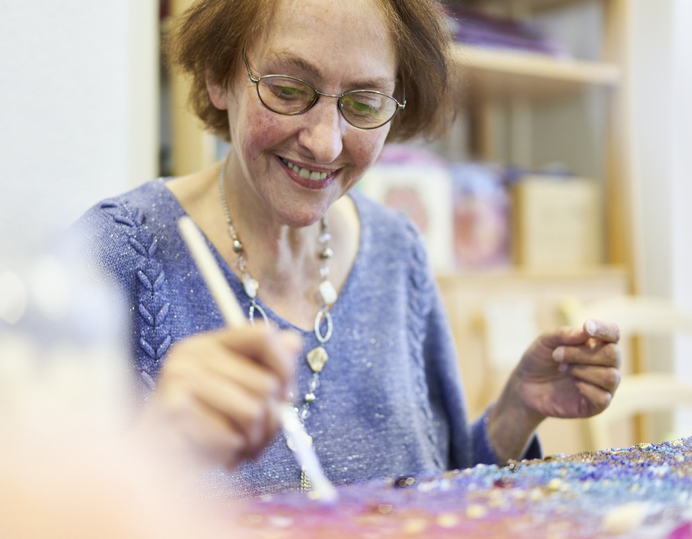 Eine ältere Dame malt mit einem Pinsel und lächelt.