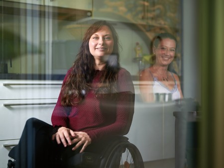 Frau im Rollstuhl lächelt in die Kamera, schräg hinter ihr steht eine andere Frau und lächelt.