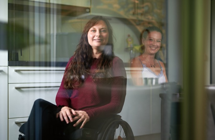 Frau im Rollstuhl lächelt in die Kamera, schräg hinter ihr steht eine andere Frau und lächelt