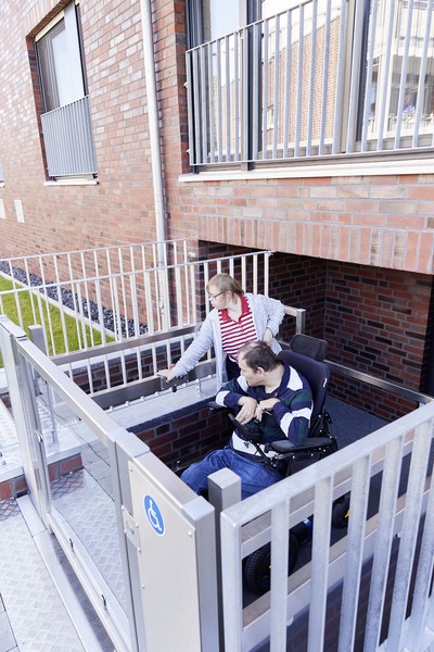 Eine junge Frau und ein Mann, der in einem elektrischen Rollstuhl sitzt, bedienen einen Aufzug, der außen an einem Gebäude ist.