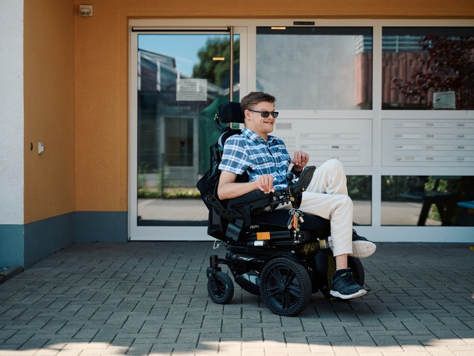 Junger Mann in einem elektrischen Rollstuhl vor einem Wohnhaus (vergrößerte Bildansicht wird geöffnet)